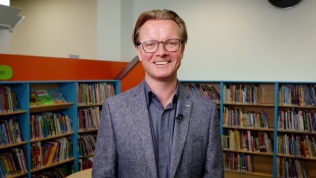 A photograph of Ed Jewell, Libraries Connected President Elect. Ed is wearing a grey jacket and is standing in front of bookshelves.