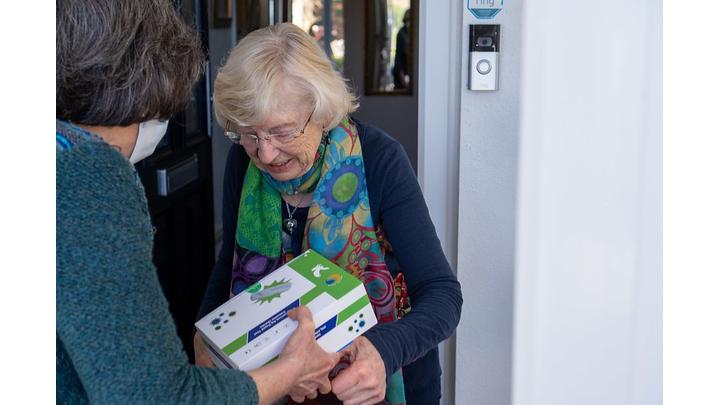 A woman hands over a box to another woman who is smiling as she receives it