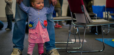 A young girl dressed in pink and blue is held up by a parent as she walks in a library