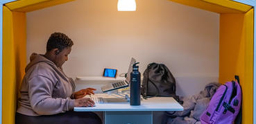 A woman with short cut black hair sits in a library pod built in the shape of a house. She is using a laptop.