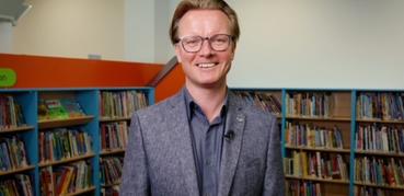A photograph of Ed Jewell, Libraries Connected President Elect. Ed is wearing a grey jacket and is standing in front of bookshelves.