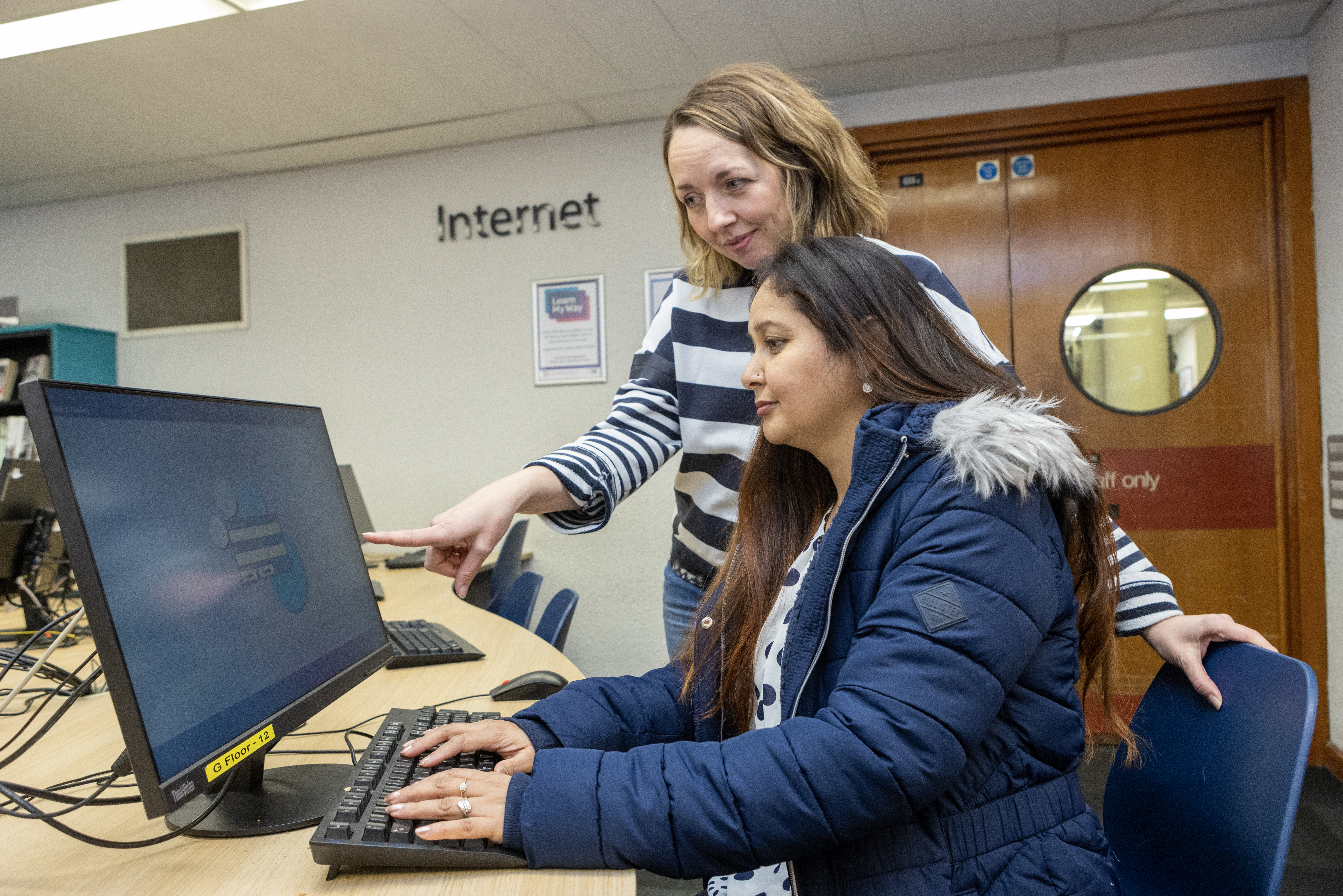 A person using a computer in a library with help from a library worker