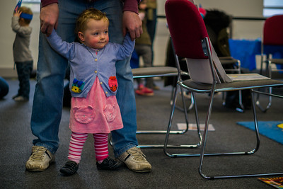A young girl dressed in pink and blue is held up by a parent as she walks in a library