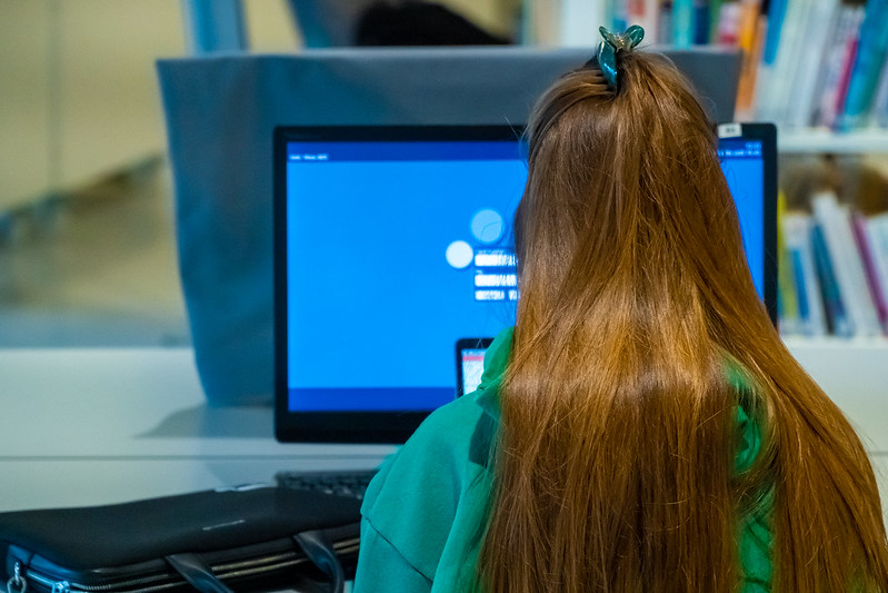 The back of a young girl's head as she works at a computer screen in a library