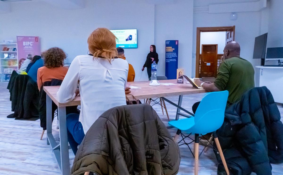 A photograph of a room in which several adults sit listening to a teacher. A photograph of a room in which several adults sit listening to a teacher.