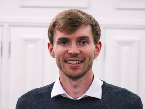 David Barclay smiles broadly into the camera. He is wearing a white collared shirt underneath a black sweater.