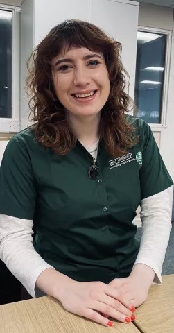 Sarah Shiels is seen smiling at the camera. Her hands are resting on a wooden surface.
