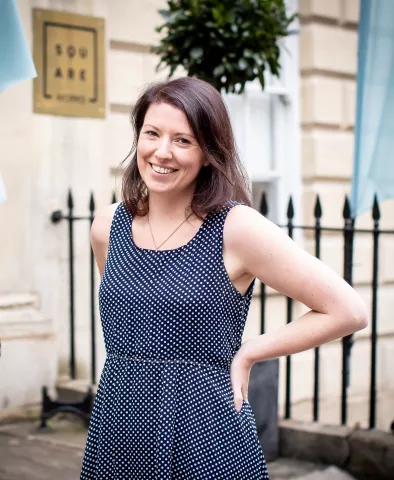 Hannah Beatrice smiles in a photograph taken in front of a pale stone building with black railings.