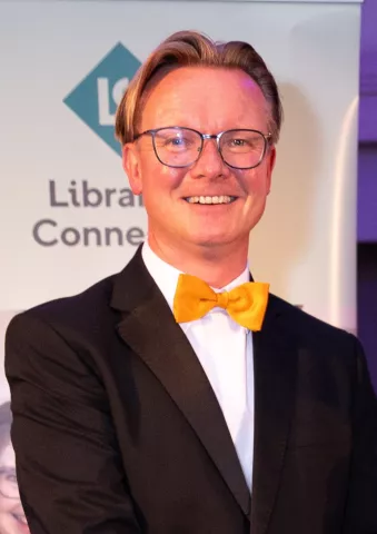Libraries Connected President Ed Jewell smiles into the camera. He is wearing a black suit with an orange bow tie.