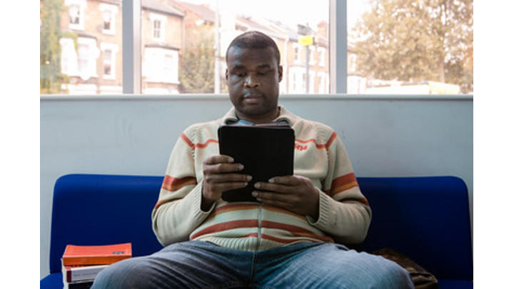 A man in a cream jumper with red stripes reads from a tablet device.