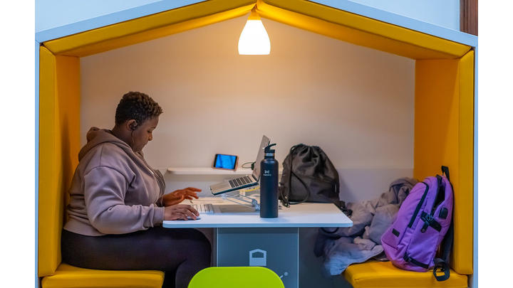 A young girl sits studying at a laptop in a library cubby hole. Above her a lamp shines.