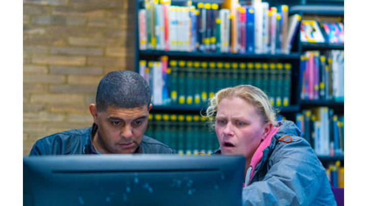 A woman wearing a coat with a pink lining helps a man use a computer. Behind them is a large bookshelf.