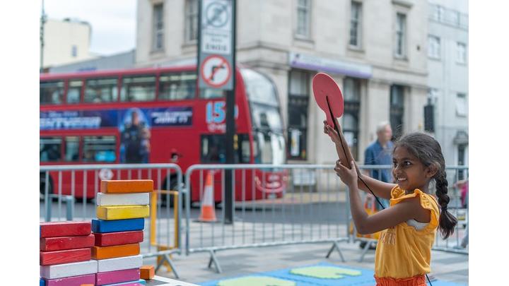 A young girl holds up a red bat with a smile on her face. In the background is a red London bus.
