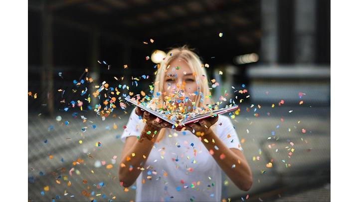 A woman with blonde hair blows confetti that scatters over an open book.