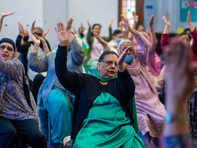  A group of older women participates in a seated exercise class, raising their arms in unison, creating a sense of community and movement.