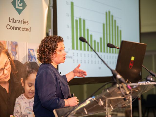 A woman is stood at a lectern giving a presentation wit a bar chart projected on to a screen behind her.