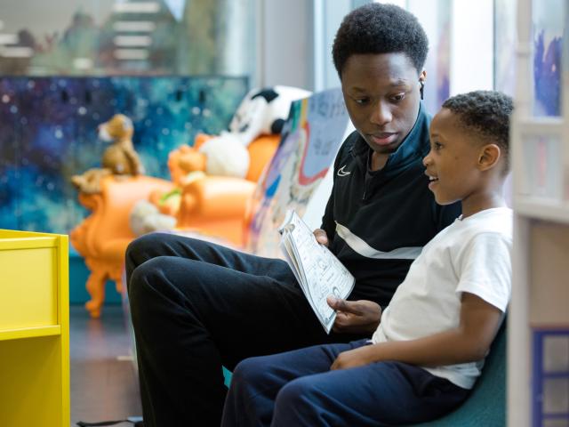 A man is sitting with a young boy, reading together in a bright, colourful children's library.