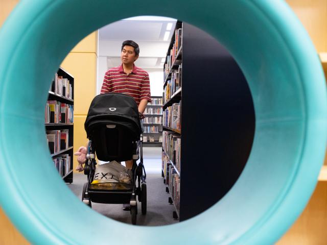 Man with a baby stroller, seen through a circular blue tube, browsing bookshelves in a library