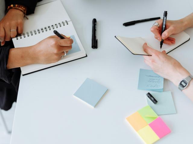 Two people writing on notepads at a white table scattered with colorful sticky notes, black markers, and notebooks