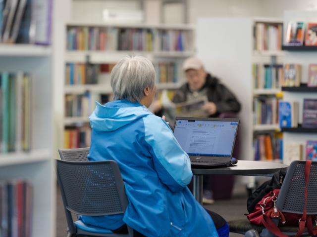 A woman sits in a library wearing a blue coat.