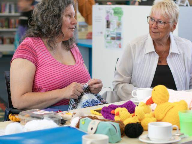 People sitting at a table in a library engaged in a craft activity with yarn and knitted items, including colourful stuffed toys. The table has cups, craft supplies and a folder. A dog is lying on the floor underneath.