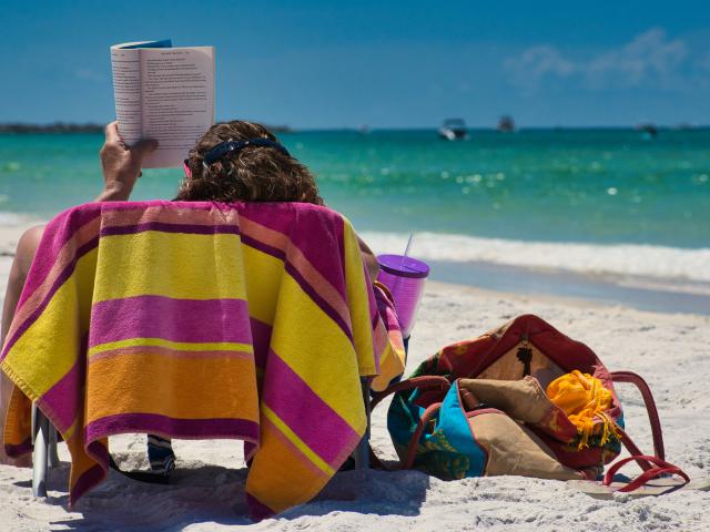 Person sitting on a sandy beach, reading a book while facing the sea, with a brightly coloured towel, a drink, and a beach bag nearby