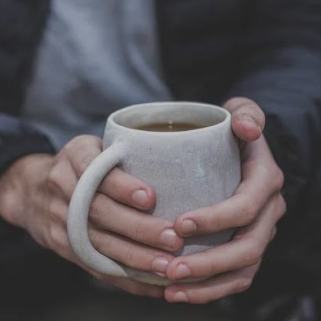 A pair of hands seen grasping a white mug of hot brown liquid.