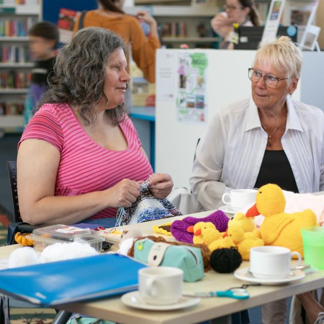 People sitting at a table in a library engaged in a craft activity with yarn and knitted items, including colourful stuffed toys. The table has cups, craft supplies and a folder. A dog is lying on the floor underneath.