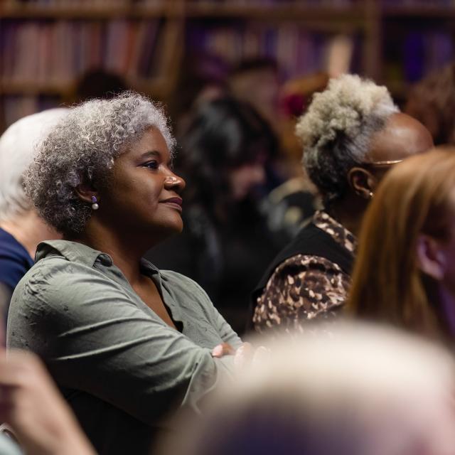 Audience seated in a library setting with bookshelves in the background, listening attentively during an event or presentation.