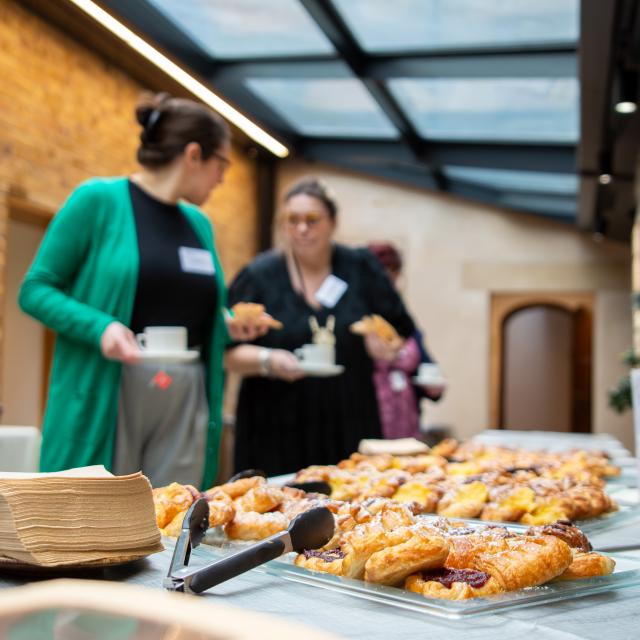 A photograph showing two women talking as they hold cups of tea. In the foreground are pastries.