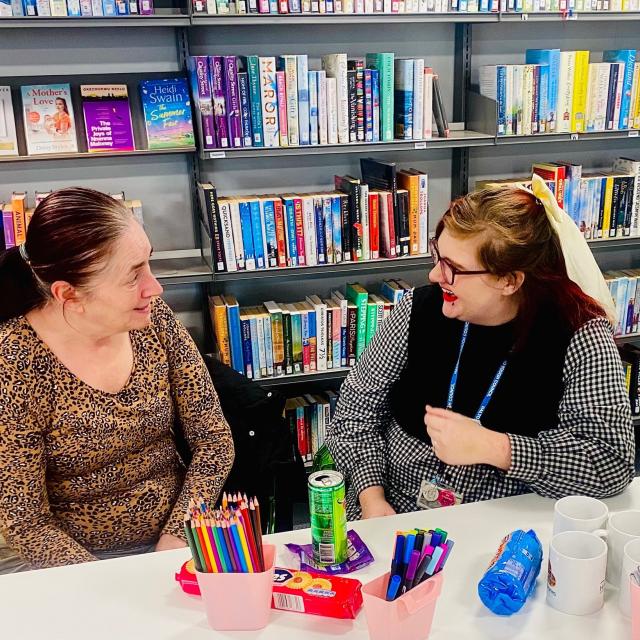 Two people sitting at a table in a library, talking, with bookshelves behind them and pens, mugs and snacks on the table.