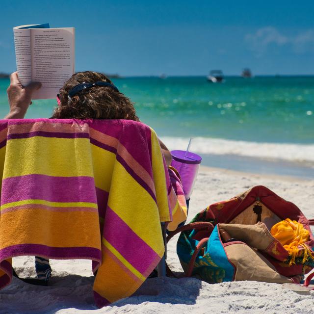 Person sitting on a sandy beach, reading a book while facing the sea, with a brightly coloured towel, a drink, and a beach bag nearby