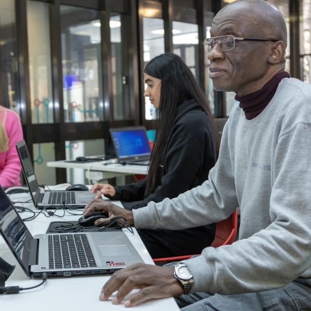 Three people sit around a table working on laptops in a bright, modern indoor space. The person in the foreground is using a laptop, with charging cables and other computer equipment spread across the table.