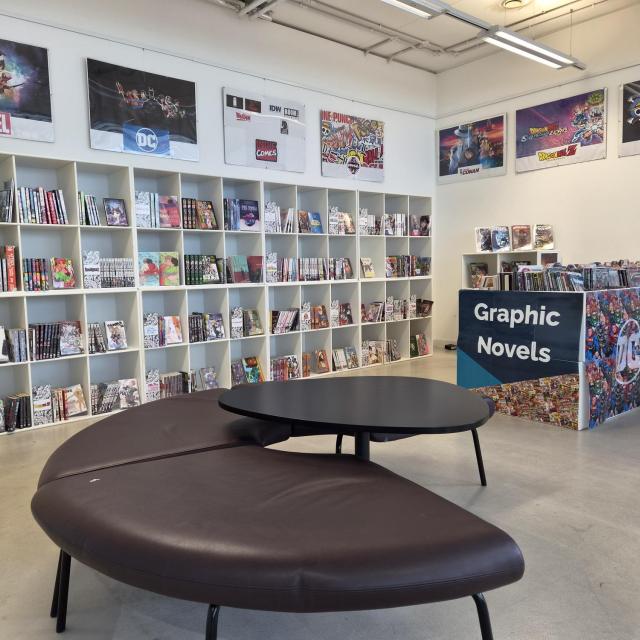 A dedicated graphic novels area in a public library, featuring wall-to-wall shelving stocked with comics, manga and graphic novels. Large posters for Marvel, DC, One Piece, Dragon Ball and other comic franchises are displayed above the shelves. In the foreground are curved seating and a low table, while a central display counter labelled “Graphic Novels” showcases additional titles. The space is bright, modern and designed for browsing and reading.