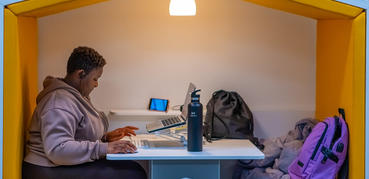 A young girl sits studying at a laptop in a library cubby hole. Above her a lamp shines.