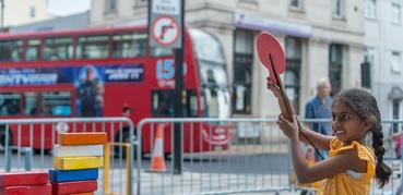 A young girl holds up a red bat with a smile on her face. In the background is a red London bus.