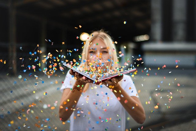 A woman with blonde hair blows confetti that scatters over an open book.
